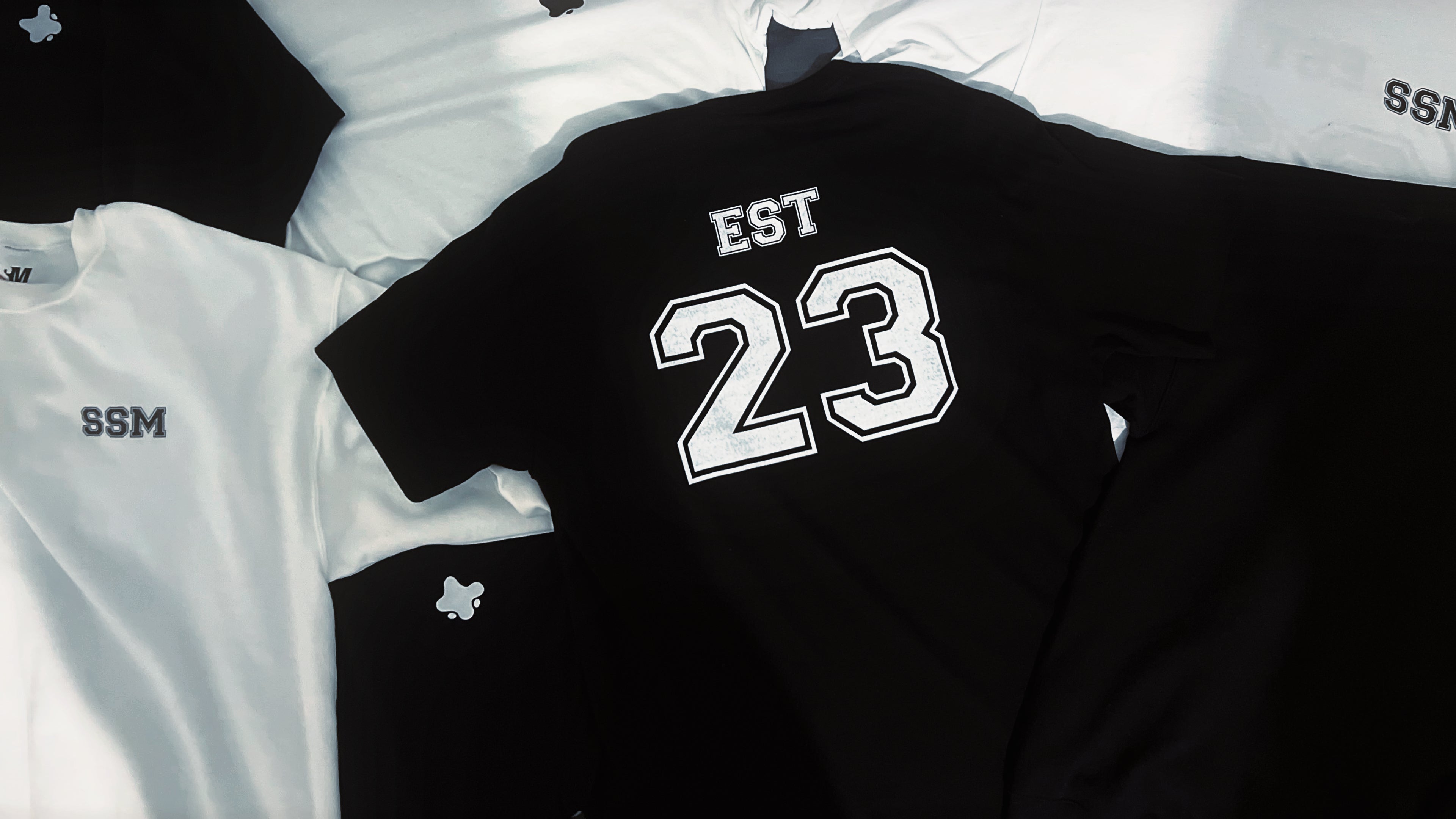 Black long-sleeve shirt with 'EST 23' printed on it, laid flat on a grey surface.