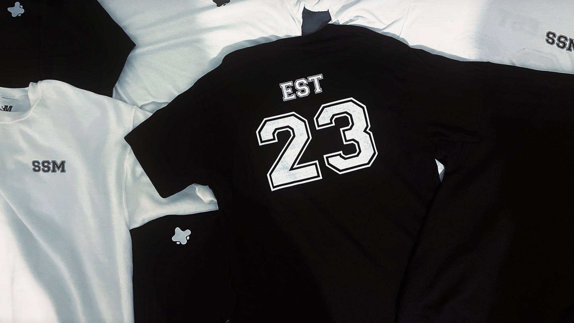 Black long-sleeve shirt with 'EST 23' printed on it, laid flat on a grey surface.